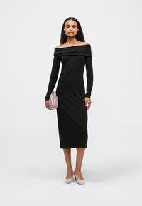 Γυναίκα φορώντας ένα μαύρο μίντι φόρεμα με μακριά μανίκια και ανοιχτούς ώμους, κρατώντας μια μικρή ροζ τσάντα και φορώντας λευκά ψηλοτάκουνα παπούτσια, στέκεται μπροστά σε λευκό φόντο.