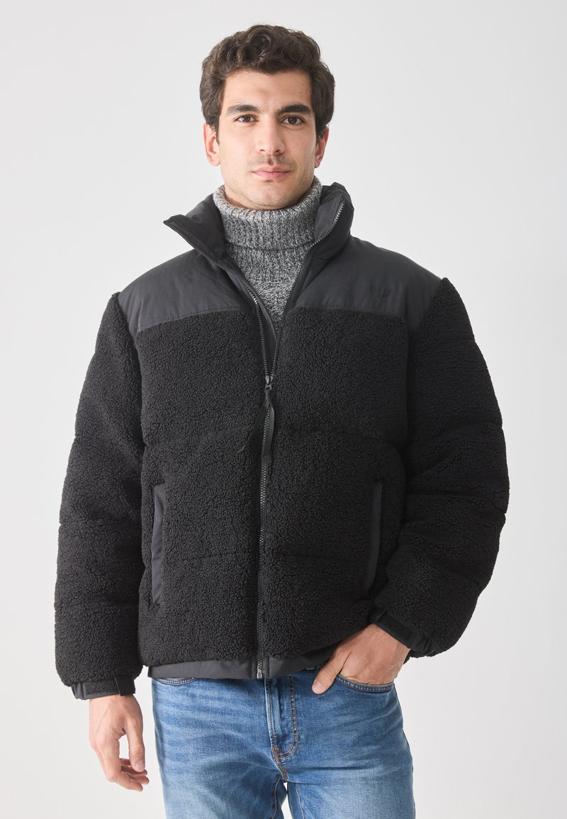 Uomo che indossa un giubbotto in pile nero sopra un maglione grigio a collo alto e jeans blu, in piedi con una mano in tasca su uno sfondo neutro.