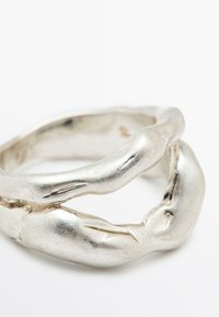 Anello d'argento con design intrecciato e testurizzato. Superficie liscia e lucida con piccole imperfezioni e bordi arrotondati, che ricordano forme organiche.