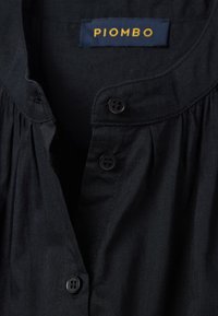 Schwarzes Hemd mit Knopfleiste, Kragen, gefalteten Details und zwei schwarzen Knöpfen. Das Etikett zeigt "PIOMBO" in Gelb auf blauem Hintergrund.