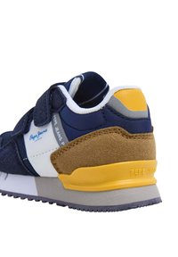 Jūras spēku sneaker ar baltām, pelēkām un dzeltenām akcentiem, ar zamša papēdi, Velcro jostu un teksturētu auduma augšu. Izturīga gumijas zole.