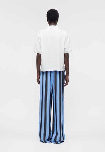 Άτομο με κοντά μαλλιά που φορά χαλαρό λευκό πουκάμισο με κοντά μανίκια και φαρδιά μπλε παντελόνια με κάθετες μαύρες ρίγες, στέκεται γυρισμένο με την πλάτη προς τα εμπρός.