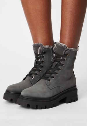 Harmaat nilkkurit, jotka on valmistettu mokasta. Niissä on pörröinen harmaa vuori, mustat nauhat ja paksut mustat kumipohjat, joissa on rosoinen kulutuspinta.