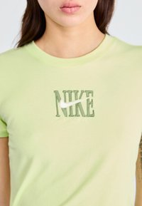 Gaiši zaļš kokvilnas T-krekls ar teksturētu "NIKE" logotipu tumšāk zaļā krāsā un baltu Swoosh, apaļa kakla izgriezums un īsas piedurknes.