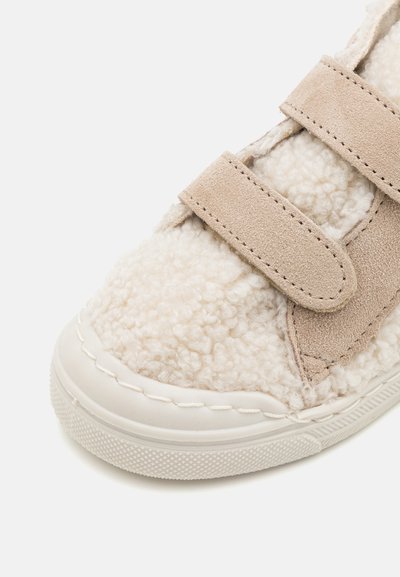 Sneaker per bambini beige con una parte superiore in tessuto soffice e due cinturini in Velcro, che si abbina a una suola in gomma bianca con un motivo testurizzato.