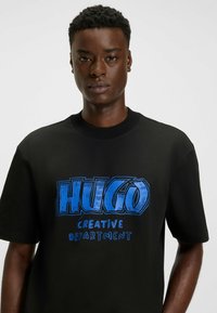Czarna bawełniana koszulka z dużą niebieską grafiką, na której widnieje napis "HUGO CREATIVE DEPARTMENT" w wyraźnej, nowoczesnej czcionce.