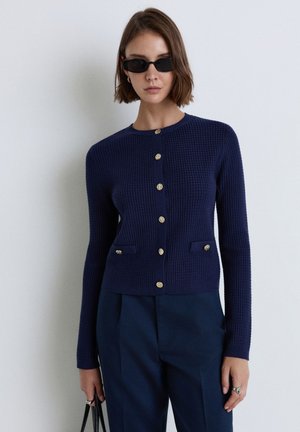 Donna che indossa occhiali da sole scuri e un cardigan di maglia blu navy con bottoni dorati, abbinato a pantaloni blu navy in tono, che tiene una borsa.