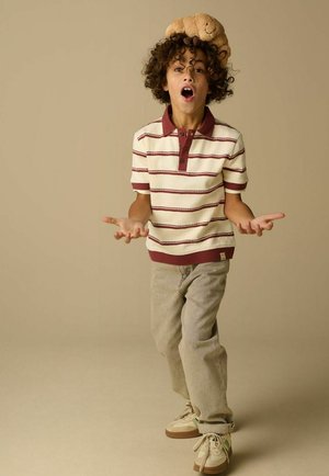 Garçon aux cheveux bouclés portant une chemise rayée et un pantalon beige, debout la bouche ouverte et les mains écartées, un ours en peluche posé sur sa tête.