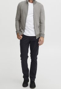 Grauer gerippter Reißverschluss-Pullover über einem weißen T-Shirt, kombiniert mit dunkelblauen Chinos und schwarzen Wildleder-Stiefeln. Sauberer, figurbetonter Schnitt und strukturierter Stoff.