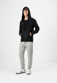 Sudadera negra con cremallera y una capucha grande, combinada con pantalones de chándal gris claro y zapatillas blancas con detalles en negro. Diseño simple y cómodo.
