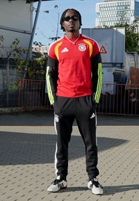 Czerwona sportowa koszulka Adidas z żółtymi i czarnymi akcentami, czarne spodnie Adidas z białymi paskami oraz czarno-białe buty. Stoi na zewnątrz.