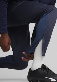 Marineblau Sporthosen mit Reißverschluss an den Knöcheln, leichtem Material und kontrastierenden blauen Seitenpanelen. Schwarze Fußballschuhe und weiße Socken sichtbar.