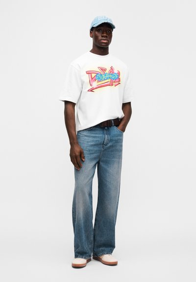 Λευκό T-shirt με πολύχρωμο γραφικό σχέδιο, σε συνδυασμό με ανοιχτό μπλε φαρδιά τζιν παντελόνια και μπεζ παπούτσια. Διαθέτει μπλε καπέλο με λογότυπο.