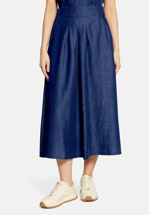 Woman wearing a high-waisted, pleated navy blue midi skirt paired with white sneakers on a plain background.