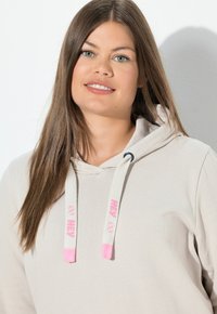 Felpa beige chiaro con cappuccio e coulisse, caratterizzata da dettagli rosa, vestibilità comoda e texture morbida. Nessun motivo o grafica visibili.