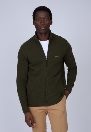 Pull à fermeture éclair vert foncé en tricot côtelé avec un col montant, accompagné d'un T-shirt blanc en dessous et d'un pantalon beige.