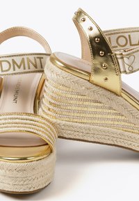 Zlatne i bijele wedge sandale s tkanim dizajnom od jute, s metalnim detaljima, remenom označenim "DMNT" i teksturiranom gornjom površinom.
