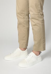 Weiße Ledersneaker mit durchbrochenen Akzenten und flachen Schnürsenkeln, kombiniert mit hellbeigen, taillierten Hosen. Einfaches, minimalistisches Design.