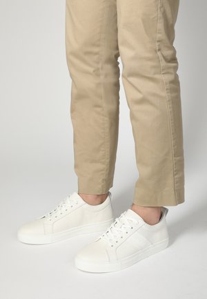 Hvide lædersneakers med perforerede detaljer og flade snørebånd, parret med lysebeige smalle bukser. Simpel, minimalistisk design.