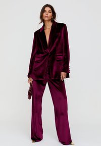 Samtiges burgunderrotes Anzugset mit einem Blazer, der einen tiefen V-Ausschnitt und zwei Vordertaschen aufweist, kombiniert mit weitem Hosenbein. Hält eine kleine Handtasche.