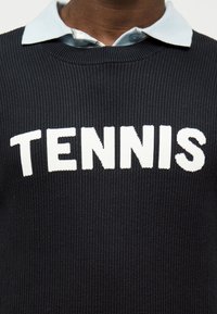 Personne portant un pull noir côtelé avec "TENNIS" en lettres blanches audacieuses sur une chemise à col bleu clair.