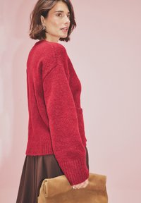 Červený pletený sveter, voľný strih s rebrovanými manžetami a lemom, nosený cez hnedú sukňu. Model drží svetlohnedý semišový clutch. Ružové pozadie.