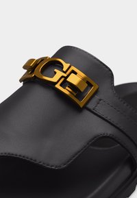 Μαύρο δερμάτινο σανδάλι slide με διακοσμητική αγκράφα σε χρυσαφένιο τόνο, σχηματισμένη από τα γράμματα «G» και «C» στο επάνω λουράκι.