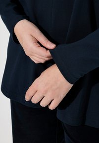 Marinblå långärmad topp med rund halsringning, i ett glatt och elastiskt tyg. Närbild på händer som justerar ärmen.