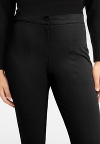 Svarta figursydda byxor i texturerat tyg, med knappstängning, bälteshällor och sidofickor. Midjebandet är slätt och elastiskt.
