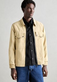Lichtbeige jack met twee borstzakken, zwarte drukknoopknopen, ritssluiting aan de voorkant en elastische manchetten, gelaagd over een zwart button-up shirt.