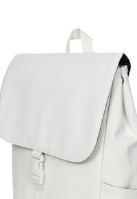Witte rugzak met een klepsluiting, gemaakt van glad materiaal. Heeft een voorste gesp en een draaghandvat. Geaccentueerd met een textuur op de randen.