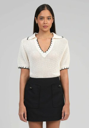 Femme aux cheveux droits et foncés porte un haut blanc en maille à manches courtes avec un col en V et des bordures noires, accompagné d'une jupe noire avec deux poches avant à boutons.