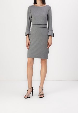 Femme portant une robe grise ajustée jusqu'aux genoux avec des manches évasées trois-quarts et des sandales à talons noirs à lanières croisées.