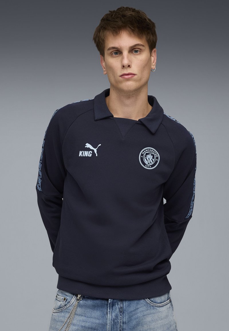 Felpa polo a maniche lunghe blu navy con colletto, con logo Puma bianco, emblema del Manchester City e accenti blu decorati sulle maniche.