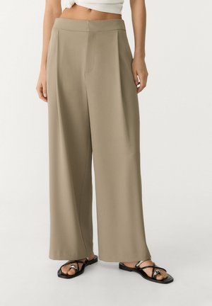 Femme portant un pantalon large beige et des sandales plates noires, debout devant un fond blanc.