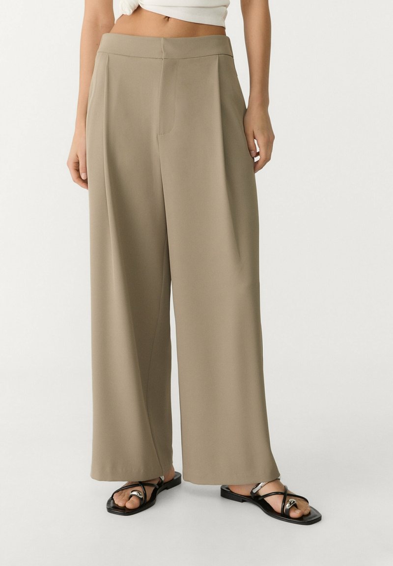 Femme portant un pantalon large beige et des sandales plates noires, debout devant un fond blanc.