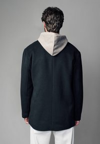 Schwarzer Wollmantel mit grauem Kapuzen, der eine schlichte Silhouette und eine glatte Textur aufweist. Die Rückseite zeigt klare Linien und keine sichtbaren Verschlüsse.