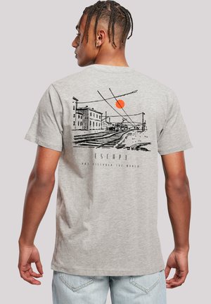 Mann mit geflochtenem Haar, der ein hellgraues T-Shirt trägt, das auf der Rückseite eine schwarze Zeichnung von Gleisen, Gebäuden und einer orangefarbenen Sonne zeigt.