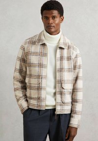 Mand iført en beige ternet zip-up jakke over en hvid turtleneck sweater og mørke bukser, der står foran en ensfarvet grå baggrund.
