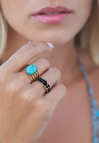 Des bagues en or avec une pierre ovale turquoise et un anneau noir comportant un motif doré, portées sur une main à la peau claire et aux ongles manucurés.