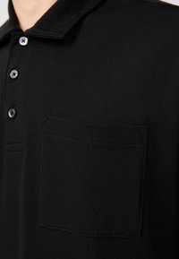 Sort polo-shirt lavet af tekstureret stof, med krave, seks knapper og en enkelt brystlomme med pæne syninger.