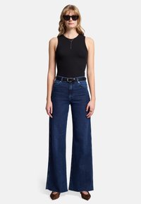 Czarny prążkowany top bez rękawów, wysokie, ciemnoniebieskie szerokie spodnie z paskiem i ciemne okulary przeciwsłoneczne. Widoczne brązowe buty w wzory.