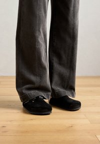 Person wearing loose, dark gray pants and black slip-on shoes standing on light wooden floor against plain wall.