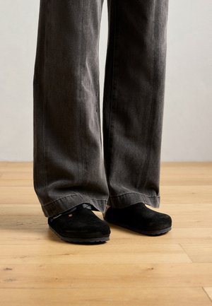 Asmuo vilkintis laisvus tamsiai pilkus kelnes ir juodus slip-on batus stovi ant šviesių medinių grindų prieš paprastą sieną.