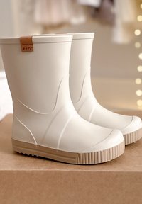 Paire de bottes de pluie en caoutchouc couleur crème avec semelles beige posées sur une surface en carton sous un éclairage intérieur doux.