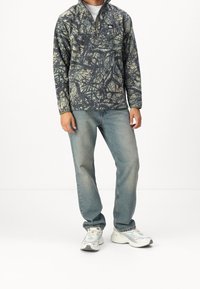 Fleece vetoketjupullovari, jossa on maastokuvio tummanvihreänä ja beigenä, yhdistettynä vaaleansinisisiin farkkuihin ja valkoisiin tennareihin.