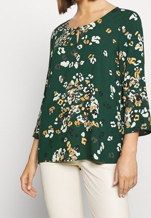 Kobieta ubrana w ciemnozieloną bluzkę z wielokolorowym kwiatowym nadrukiem i rękawami o długości trzy czwarte w kształcie dzwonka, w połączeniu z jasnobeżowymi spodniami.