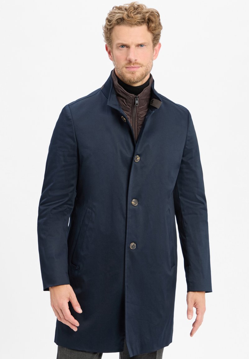 Man met krullend blond haar, gekleed in een marineblauwe jas over een bruin gewatteerd zip-up jack, staande tegen een witte achtergrond.