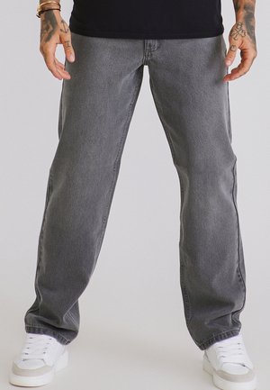 Persona che indossa jeans grigi larghi e sneakers bianche, in piedi con le mani rilassate ai lati davanti a uno sfondo semplice.