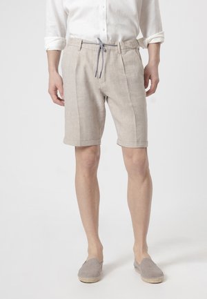 Hombre con pantalones cortos de lino beige con cordón a rayas, camisa blanca remangada y zapatos tipo slip-on grises sobre un fondo liso.